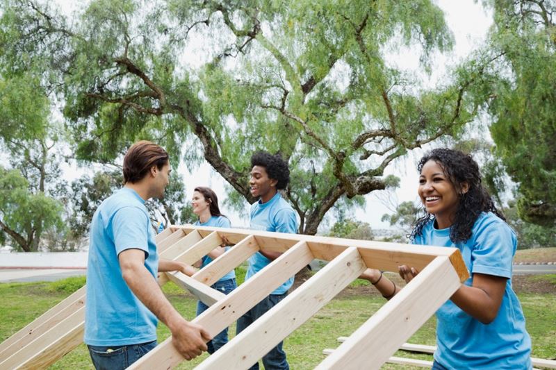 A group of volunteers wearing blue shirts are lifting the wooden frame of a wall.